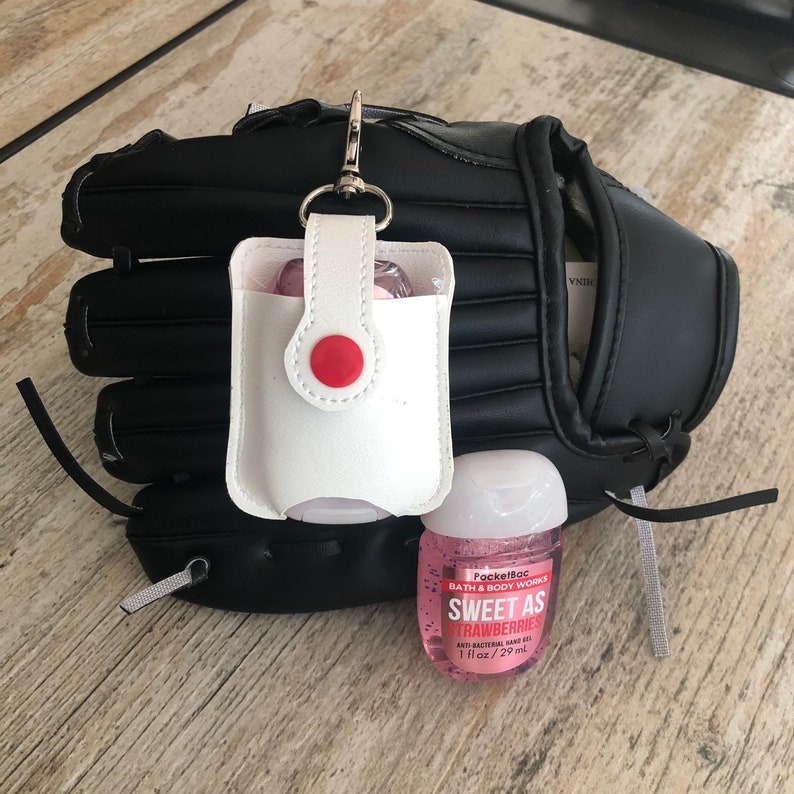 May include: A white leather hand sanitizer holder with a red button attached to a black baseball glove. A pink bottle of PocketBac Sweet as Strawberries anti-bacterial hand gel is next to the holder. The bottle is 1 fl oz / 29 ml.