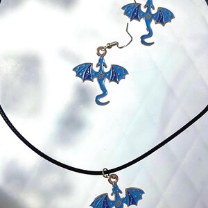 May include: A set of jewelry consisting of a black cord necklace and two matching earrings, each featuring a blue enamel dragon pendant with gold-tone sun and star details on the wings. The dragon charms are suspended from standard metal hooks.