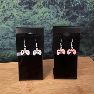 May include: Two pairs of dangle earrings featuring miniature video game controller charms. One pair is white and the other is light pink. Each charm has a small red heart in the center, colorful buttons, and silver-toned metal earring hooks.