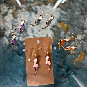 Puede incluir: Una colección de pendientes colgantes con forma de gato y ganchos dorados. Los pendientes presentan diseños de esmalte en varias combinaciones de colores, incluyendo morado, negro, blanco, naranja y rosa, sobre una tarjeta marrón.