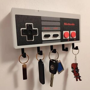 Puede incluir: Un llavero de pared diseñado para parecerse a un mando de juego clásico de Nintendo. La unidad rectangular presenta una cara negra y gris con un pad direccional, botones Select y Start, y dos botones rojos A y B. Cinco ganchos negros sostienen varias llaves.