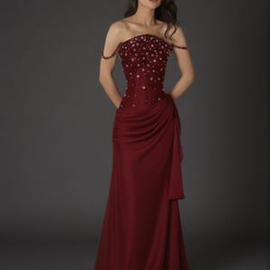 May include: A burgundy strapless evening gown with a fitted bodice and a flowing skirt. The bodice is embellished with small, scattered beadwork. The dress features a draped detail on the side and a subtle train. The model is wearing the dress.