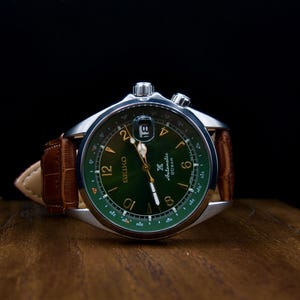 Könnte beinhalten: Nahaufnahme einer Seiko-Uhr mit grünem Zifferblatt und braunem Lederarmband. Die Uhr hat ein silbernes Gehäuse und ein Datumsfenster. Die Uhr liegt auf einer Holzoberfläche. Das Zifferblatt misst ca. 4 cm im Durchmesser.