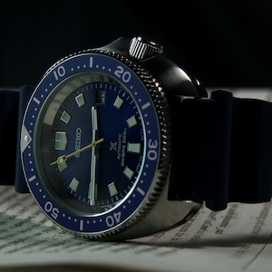 Könnte beinhalten: Nahaufnahme einer Seiko Automatik-Taucheruhr mit blauem Zifferblatt und Lünette. Die Uhr hat ein schwarzes Kautschukarmband und ein silbernes Gehäuse. Das Zifferblatt zeigt die Zeit mit weißen Markierungen und Zeigern an.