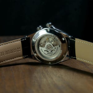 May include: Close-up of a Seiko watch, showcasing the intricate inner workings through a transparent back. The watch features a black leather strap with "GENUINE LEATHER" text. The watch is on a wooden surface.
