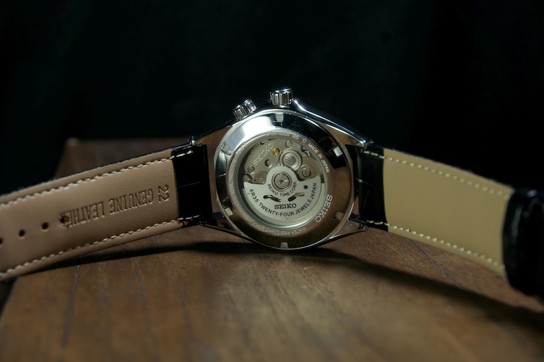 May include: Close-up of a Seiko watch, showcasing the intricate inner workings through a transparent back. The watch features a black leather strap with "GENUINE LEATHER" text. The watch is on a wooden surface.