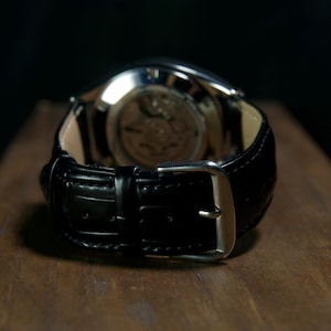 May include: Close-up of a black leather watch with a silver buckle. The watch's back is visible, showcasing the intricate inner workings. The watch is set on a wooden surface, with a dark background.