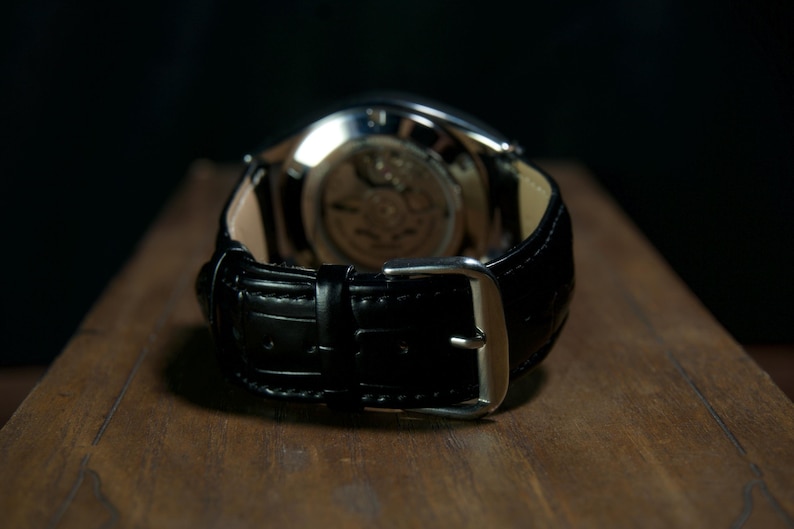 May include: Close-up of a black leather watch with a silver buckle. The watch's back is visible, showcasing the intricate inner workings. The watch is set on a wooden surface, with a dark background.