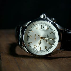 May include: A silver-toned Seiko watch with a light-colored face and black leather band. The watch face features gold-colored hands and numerals, with a date display at the 3 o'clock position. The watch is set against a dark background.