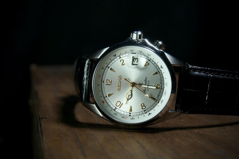 May include: A silver-toned Seiko watch with a light-colored face and black leather band. The watch face features gold-colored hands and numerals, with a date display at the 3 o'clock position. The watch is set against a dark background.