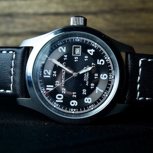 Op de afbeelding: Een close-up van een zwart wijzerplaat horloge met een zilveren kast en een zwarte leren band met witte stiksels. De wijzerplaat geeft de tijd en datum weer. Het horloge ligt op een donkere ondergrond.
