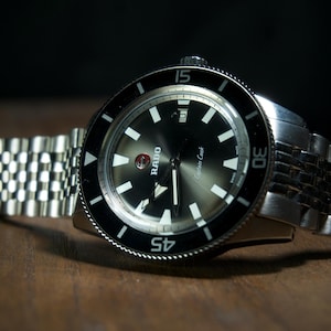 May include: A close-up of a silver wristwatch with a black dial and a black rotating bezel. The watch has white hour markers and hands. The watch band is made of silver metal links. The watch is resting on a wooden surface.