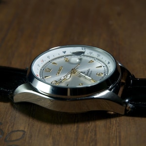May include: A silver-toned wristwatch with a black leather band. The watch face is silver with gold-colored hands and numerals. The watch is set against a dark wooden surface.