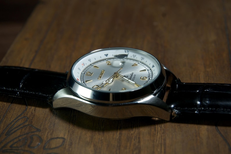May include: A silver-toned wristwatch with a black leather band. The watch face is silver with gold-colored hands and numerals. The watch is set against a dark wooden surface.