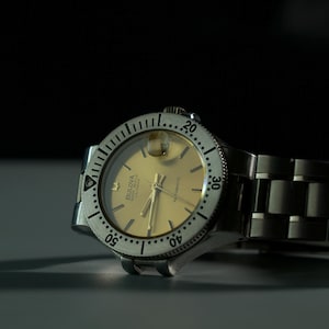 Op de afbeelding: Een zilverkleurig Bulova automatisch polshorloge met een goudkleurige wijzerplaat. Het horloge heeft een draaibare bezel met minutenmarkeringen en een roestvrijstalen band. Het horloge staat tegen een donkere achtergrond.