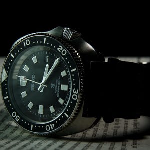 Pode incluir: Um relógio de mergulho automático Seiko preto com mostrador preto, marcadores luminosos e moldura rotativa. O relógio tem uma bracelete de borracha preta e é exibido numa superfície texturizada.