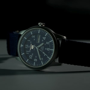 May include: A dark blue analog watch with a black strap. The watch face is dark blue with white numerals and hands. The watch has a day and date display. The watch is set against a dark background.