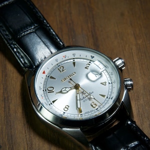 May include: A silver-faced Seiko watch with a black leather band. The watch has a white dial with black numerals and hands, and a date window. The watch is set against a wooden surface.