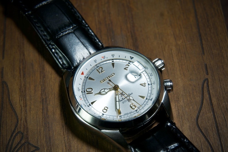 May include: A silver-faced Seiko watch with a black leather band. The watch has a white dial with black numerals and hands, and a date window. The watch is set against a wooden surface.