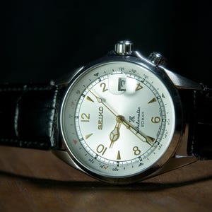 May include: A silver-toned Seiko watch with a light-colored face and black leather band. The watch face displays the Seiko logo, numerals, and hands. The watch has a date window and two crowns. The band is textured.