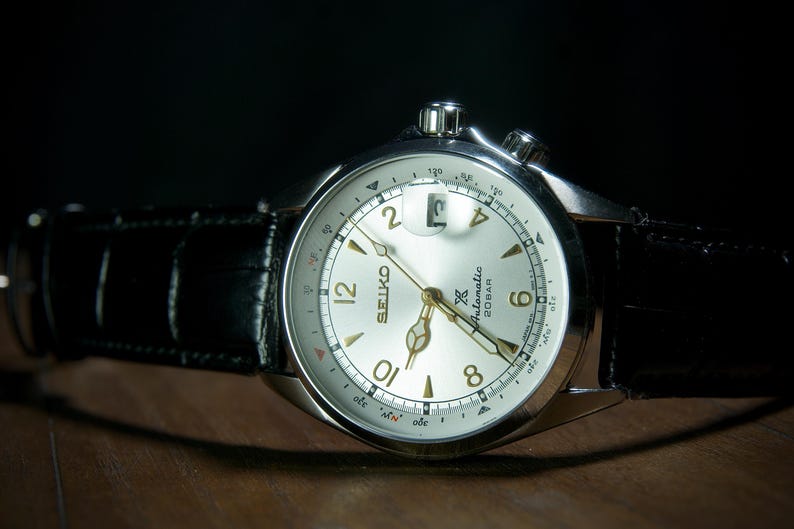 May include: A silver-toned Seiko watch with a light-colored face and black leather band. The watch face displays the Seiko logo, numerals, and hands. The watch has a date window and two crowns. The band is textured.