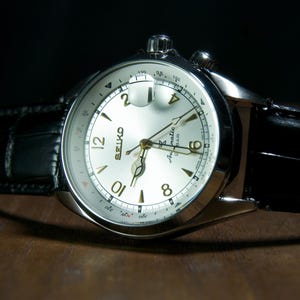 May include: A close-up of a silver-toned Seiko watch with a white face and gold-colored hands and numerals. The watch has a black leather band. The watch is on a wooden surface with a dark background.