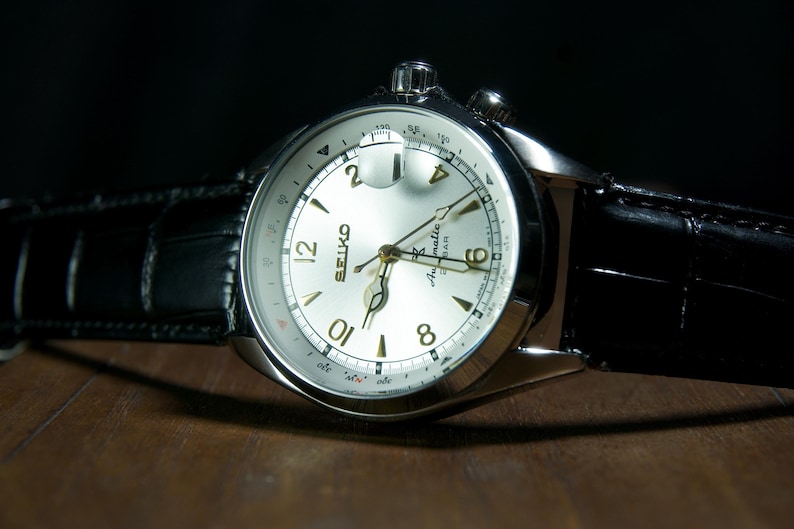 May include: A close-up of a silver-toned Seiko watch with a white face and gold-colored hands and numerals. The watch has a black leather band. The watch is on a wooden surface with a dark background.