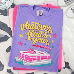 Puede incluir: Tres camisetas apiladas en tonos de rosa y morado. La camiseta superior es morada con el texto "whatever floats your" en escritura amarilla, con un gráfico de barco pontón rosa. Las otras camisetas son de color rosa liso.