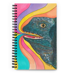 May include: Spiral-bound notebook featuring a colorful illustration of a reptile's head with an open mouth. The artwork includes a teal reptile with an orange eye, set against a vibrant rainbow background of pink, purple, yellow, and orange.