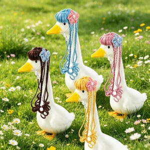 Puede incluir: Cuatro figuras de gansos blancos con picos y patas amarillas, cada una adornada con cabello de hilo de diferentes colores y una flor. Los gansos están en un área cubierta de hierba con pequeñas flores blancas y amarillas.