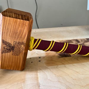 Puede incluir: Un mazo de madera con una cabeza marrón claro y un mango envuelto en cordón burdeos y amarillo. La cabeza del mazo presenta un diseño grabado de águila y galones. El mango mide unos 25 cm.