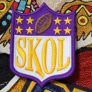 May include: Embroidered patch with a purple shield design, featuring a brown football, gold stars, and the word "SKOL" in gold lettering. The patch is surrounded by other colorful embroidered patches. The patch is likely for a sports fan.