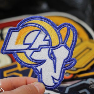 May include: Embroidered patch featuring the Los Angeles Rams logo, a ram's head with the letters "LA" in yellow and white, outlined in blue. The patch is held in front of a blurred background of other patches.