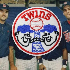 May include: Image of three baseball players in navy blue uniforms, holding bats. The players are standing in front of a large Twins logo patch. The patch is circular, with the word "TWINS" in red and a baseball in the center.