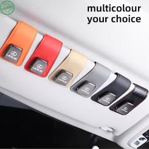 Puede incluir: Seis soportes para gafas de visera de coche en naranja, rojo, beige, gris, negro y patrón de fibra de carbono, cada uno con un clip metálico plateado con el logo de Lexus. El texto multicolour your choice está impreso en la superficie gris.