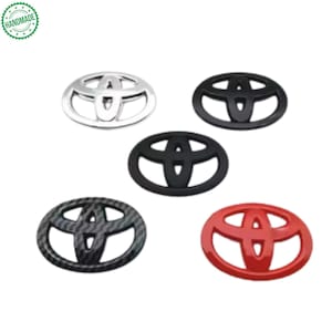 May include: Five car emblem badges in various colors and finishes. The badges include silver, black, carbon fiber, and red. The Toyota logo is visible on each badge. The word "HANDMADE" is printed on a green circular sticker in the upper left corner.