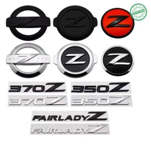 May include: A collection of car emblems and badges, including black, silver, and red designs. The emblems feature the letter "Z" and text such as "370Z", "350Z", and "Fairlady Z". Some are circular, while others are rectangular.