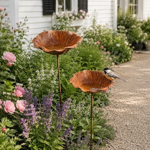 May include: Two copper-colored metal bird baths shaped like large, textured flower petals, each mounted on a thin, dark metal stake. A small black, white, and gray chickadee perches on the rim of the lower bird bath, which contains water.