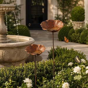 May include: Two copper-colored metal bird baths shaped like open flower blossoms, each mounted on a thin, dark metal stake. The bowls have a textured, weathered finish and are positioned at different heights among green garden foliage.