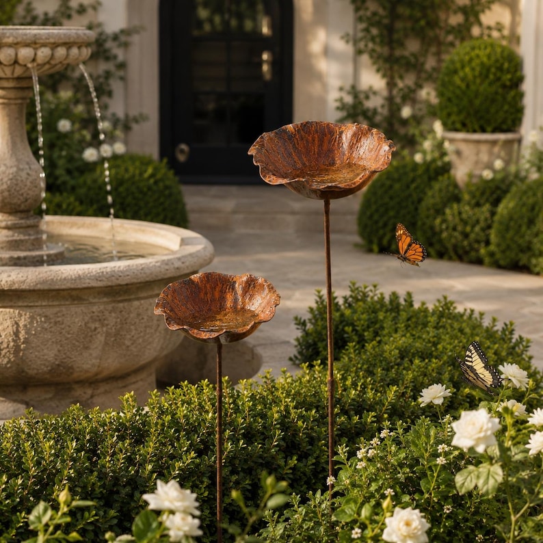 May include: Two copper-colored metal bird baths shaped like open flower blossoms, each mounted on a thin, dark metal stake. The bowls have a textured, weathered finish and are positioned at different heights among green garden foliage.