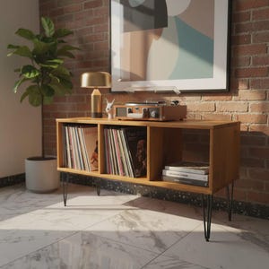 Puede incluir: Una consola de tocadiscos de madera con patas de horquilla negras. La consola tiene tres compartimentos abiertos llenos de discos de vinilo y libros. Un tocadiscos, una lámpara dorada y un pequeño conejo decorativo están encima. Una planta en maceta y una obra de arte están en el fondo.