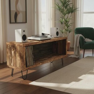 May include: A wooden record player console with black hairpin legs. The console has a built-in record storage compartment and holds a white turntable and two white speakers. A green armchair and a houseplant are in the background.