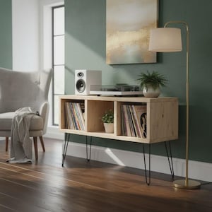 May include: A light-coloured wooden record console with black hairpin legs. The console has three open compartments filled with vinyl records and a small potted plant. A white speaker and turntable sit on top. A gold floor lamp and a grey armchair are in the background.