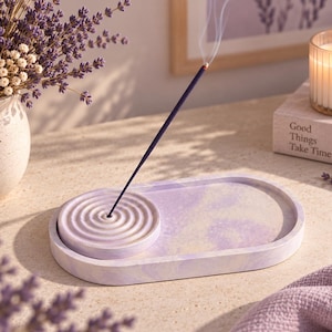 May include: An oval-shaped lavender and white marbled incense tray featuring a circular, raised platform with concentric rings for holding a single incense stick. The tray has a smooth, matte finish and a light purple and white swirled pattern.