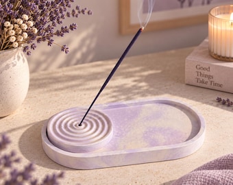May include: An oval-shaped lavender and white marbled incense tray featuring a circular, raised platform with concentric rings for holding a single incense stick. The tray has a smooth, matte finish and a light purple and white swirled pattern.