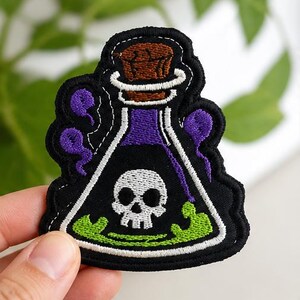 May include: Embroidered patch of a black potion bottle with a skull, purple liquid, and green bubbles. The bottle has a brown cork and white stitching. A Halloween-themed accessory.