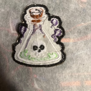 May include: Embroidered patch depicting a potion bottle. The white bottle contains a skull and green liquid. The patch has a black border and purple details, suitable for customising garments or accessories.