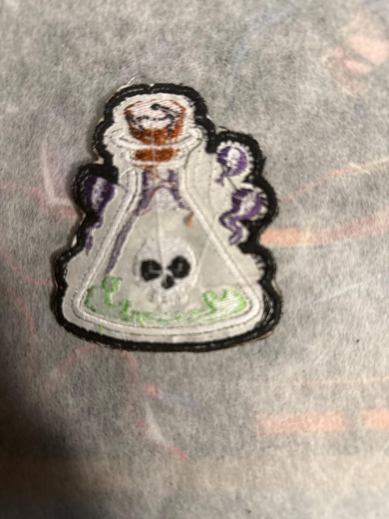 May include: Embroidered patch depicting a potion bottle. The white bottle contains a skull and green liquid. The patch has a black border and purple details, suitable for customising garments or accessories.