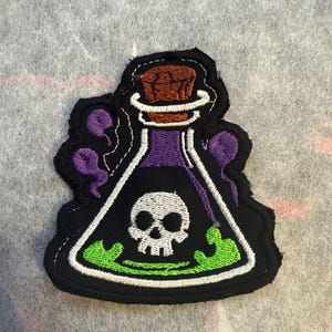 May include: Embroidered patch with a potion bottle design. The bottle is black with a white skull, purple liquid, and green bubbling liquid. It has a brown cork and purple accents.