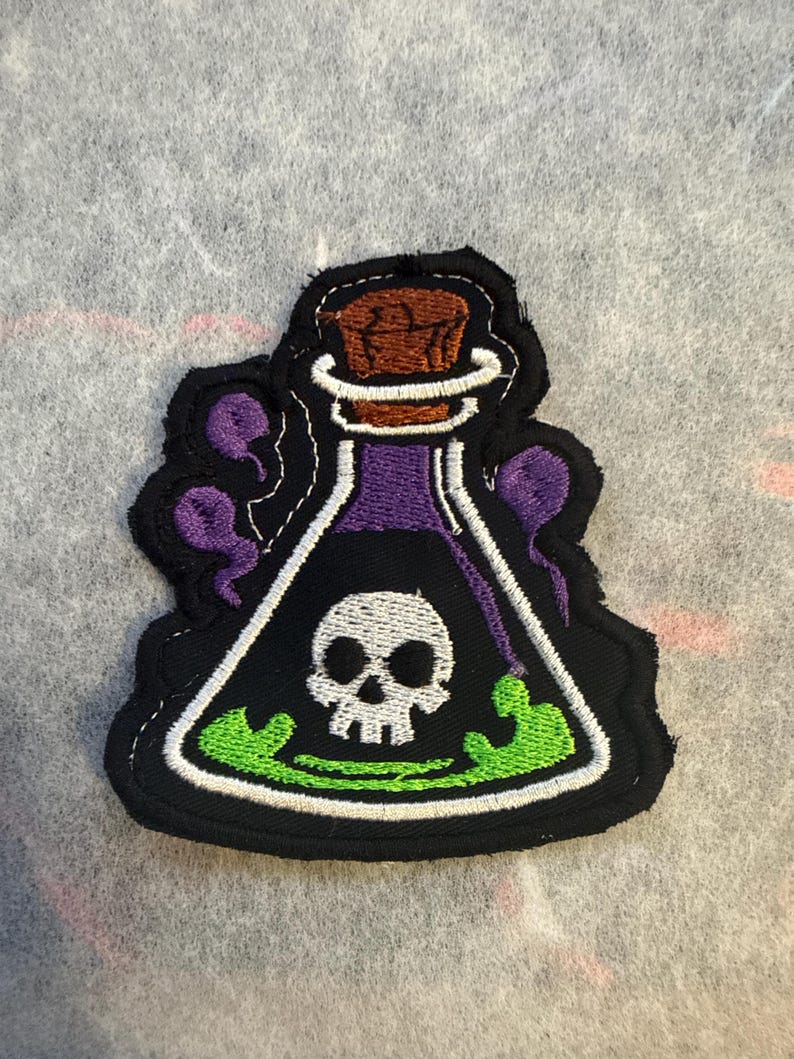 May include: Embroidered patch with a potion bottle design. The bottle is black with a white skull, purple liquid, and green bubbling liquid. It has a brown cork and purple accents.